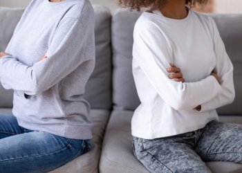 Close up African American mother and teen daughter after quarrel, sitting together on couch at home, ignore each other, stubborn mum and teenage child upset by agreement, family conflict concept (Getty Images)