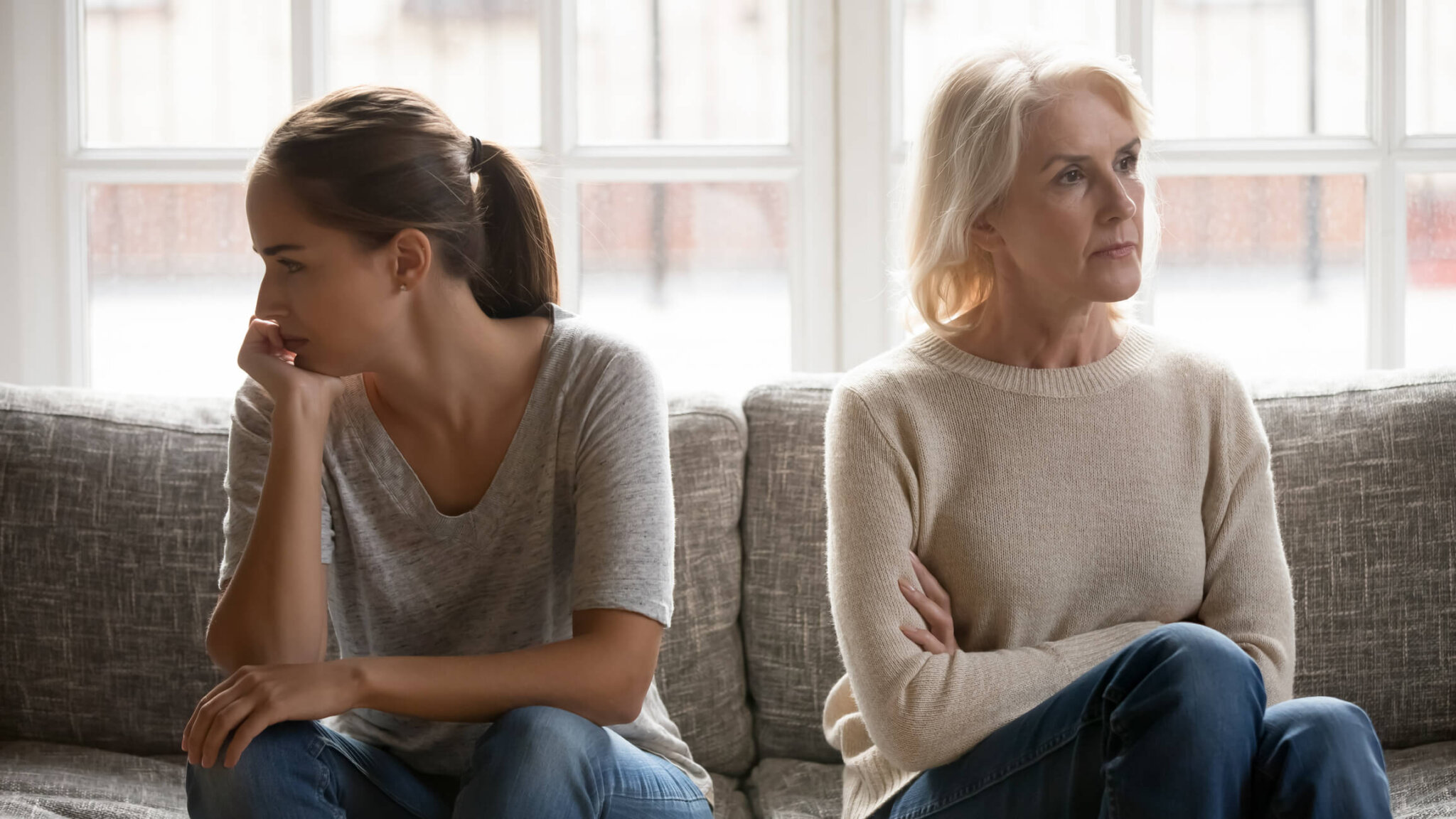 In quarrel elderly mother grown up daughter sit on couch separately having conflict, emotionally intergenerational misunderstanding, adult grandchild grandma difficult bad relations different generations concept