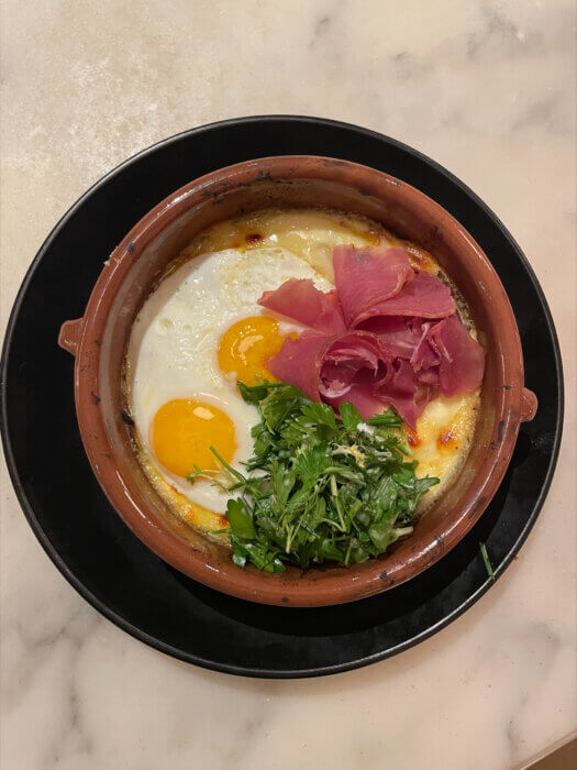 Brunch at Lulu Kitchen includes a Raclette Fonduta for two with two sunny-side-up eggs, prosciutto, Raclette cheese, potato, pickled cucumber, and truffled herb salad.