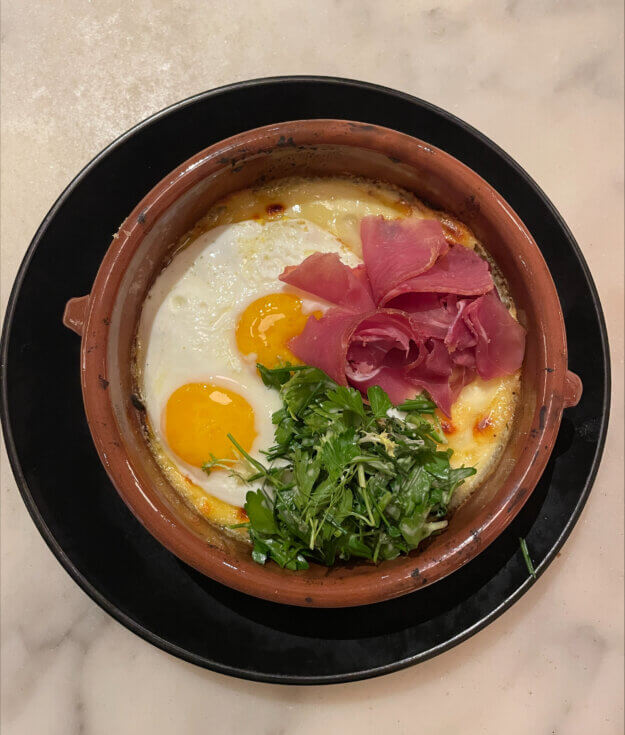 Brunch at Lulu Kitchen includes a Raclette Fonduta for two with two sunny-side-up eggs, prosciutto, Raclette cheese, potato, pickled cucumber, and truffled herb salad.