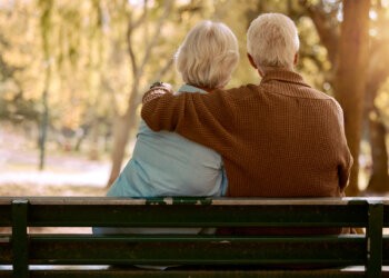 Love, hug and old couple in a park on a bench for a calm, peaceful or romantic summer marriage anniversary date. Nature, romance or back view of old woman and elderly partner in a relaxing embrace Golden Bachelor