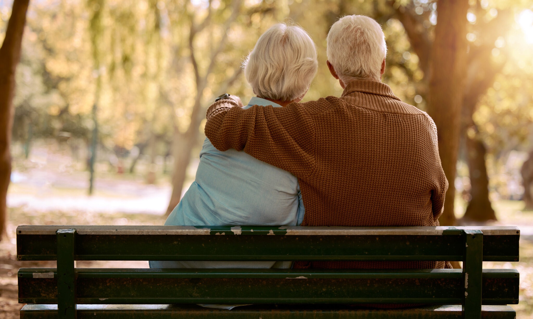 Love, hug and old couple in a park on a bench for a calm, peaceful or romantic summer marriage anniversary date. Nature, romance or back view of old woman and elderly partner in a relaxing embrace Golden Bachelor