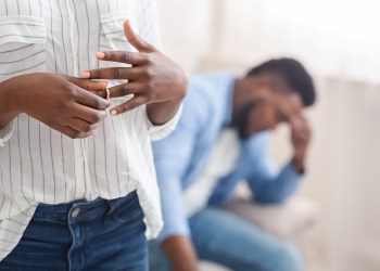 When do you leave your relationship? Divorce concept. Unrecognizable black woman removing wedding ring from finger, her despaired husband on background