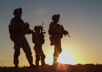 Squad of Three Fully Equipped and Armed Soldiers Standing on Hill in Desert Environment in Sunset Light. War