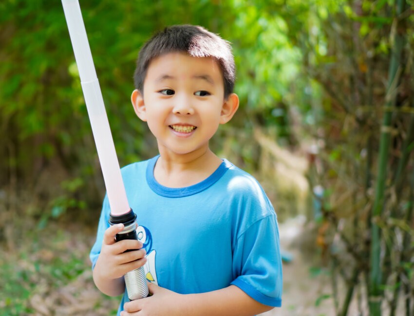 The picture shows a boy smiling happily as he plays a kids lightsaber duel with his brother.