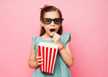 Adorable elementary age kids enjoying eating popcorn while watching a 3d movie with glasses in a pink background
