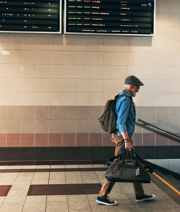 Stylish elderly man walking through train station, traveling home, holding duffel bag. Senior man entering the subway station.