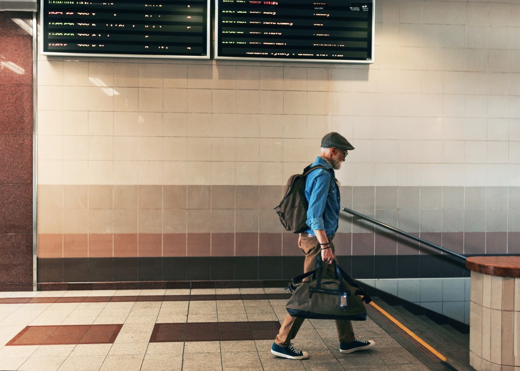 Stylish elderly man walking through train station, traveling home, holding duffel bag. Senior man entering the subway station.