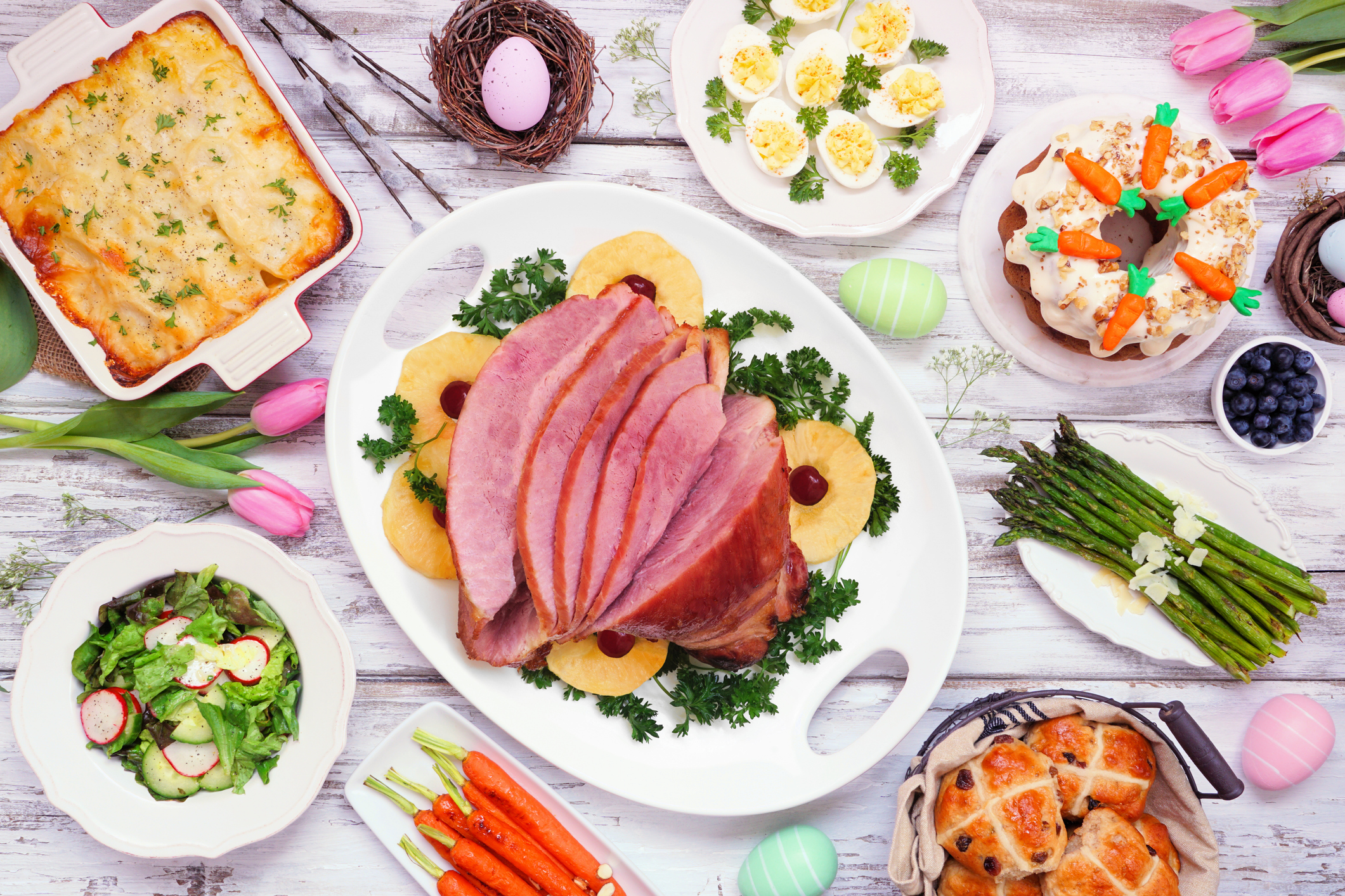 Traditional Easter ham dinner. Top view table scene on a white wood background. Ham, scalloped potatoes, eggs, hot cross buns, carrot cake and vegetables.