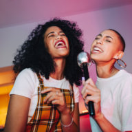 Best friends singing into a microphone on karaoke night. Two cheerful young women singing their favourite song at a house party. Happy female friends having a good time during the weekend in the Hamptons