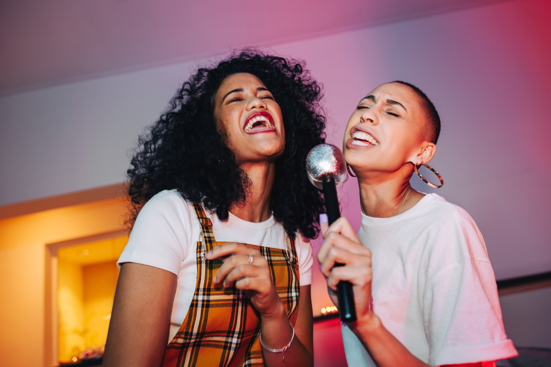 Best friends singing into a microphone on karaoke night in the Hamptons. Two cheerful young women singing their favourite song at a house party. Happy female friends having a good time during the weekend in the Hamptons