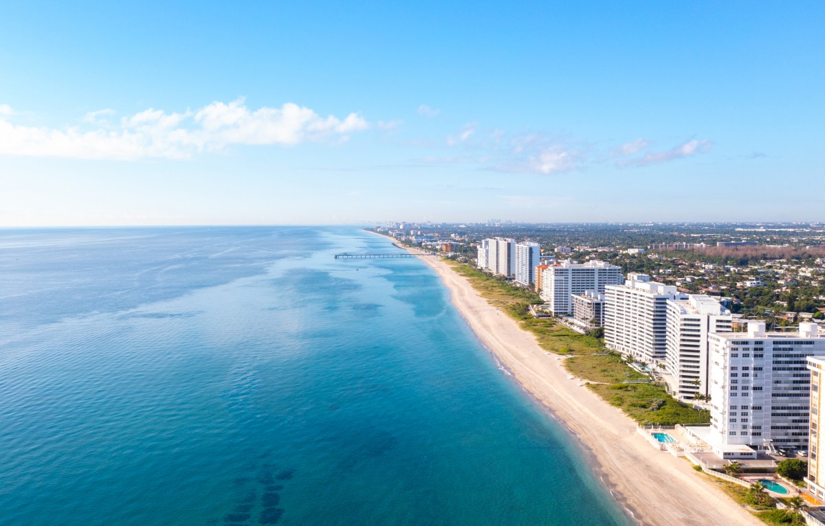 Aerial photograph of Boca Raton, Florida, showcasing the pristine Atlantic Ocean coastline, golden sandy beaches, and luxury oceanfront condominiums. The panoramic view captures the serene turquoise waters, bright blue sky, and peaceful coastal lifestyle, highlighting South Florida’s scenic beauty and upscale beachfront living
